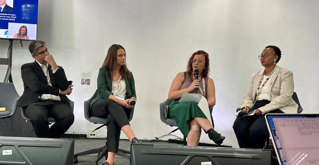 Four panelists sit on stage during a COP30 climate discussion. One speaker holds a microphone and notes while the others listen. A presentation screen and conference badges are visible in the background.